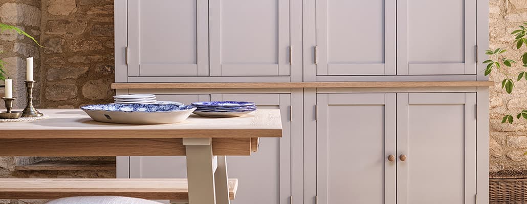 Rustic kitchen with light gray cabinets, wooden countertops, a wooden table with blue-patterned plates, and stone walls.