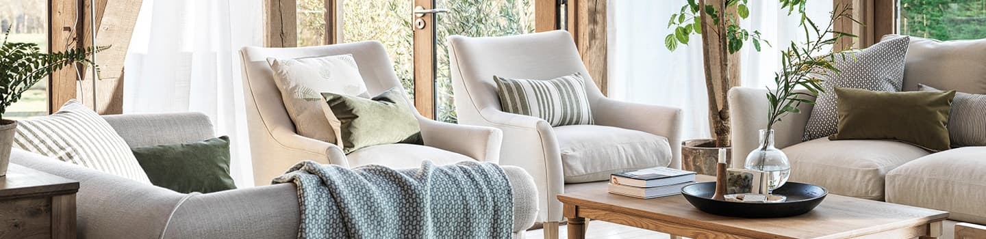 Cozy living room with beige armchairs, green and striped pillows, a wooden coffee table, and potted plants near large windows with sheer curtains.