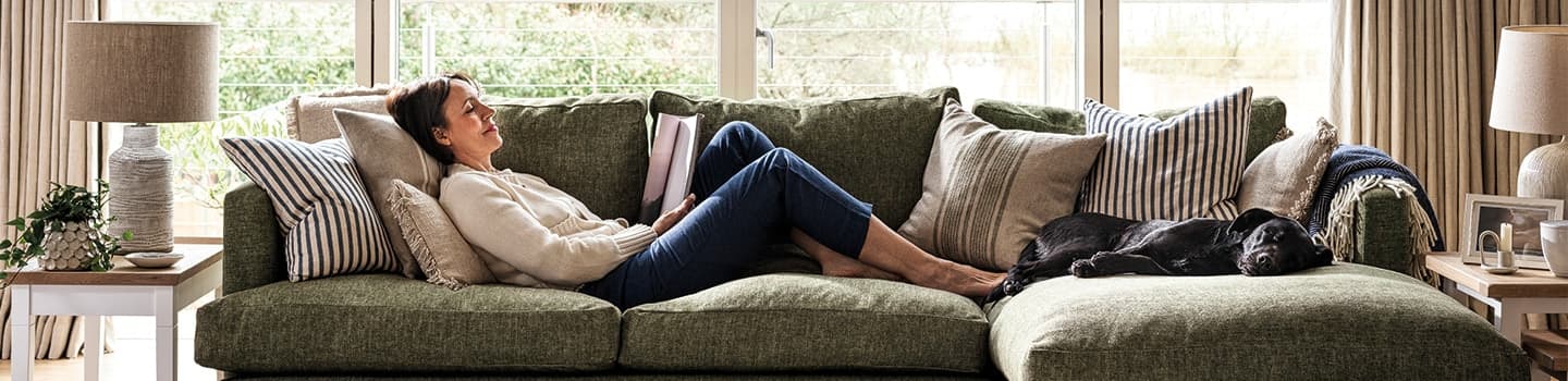 Woman relaxing on a green sofa with a black dog, surrounded by cushions, near large windows with natural light.