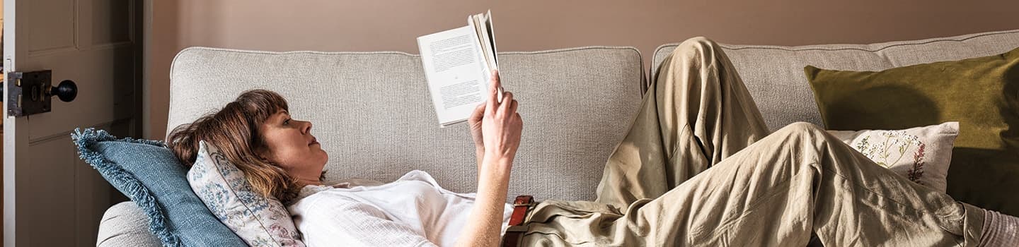 A person reclines on a sofa, reading a book. They are lying on their back with a cushion under their head, surrounded by soft pillows.