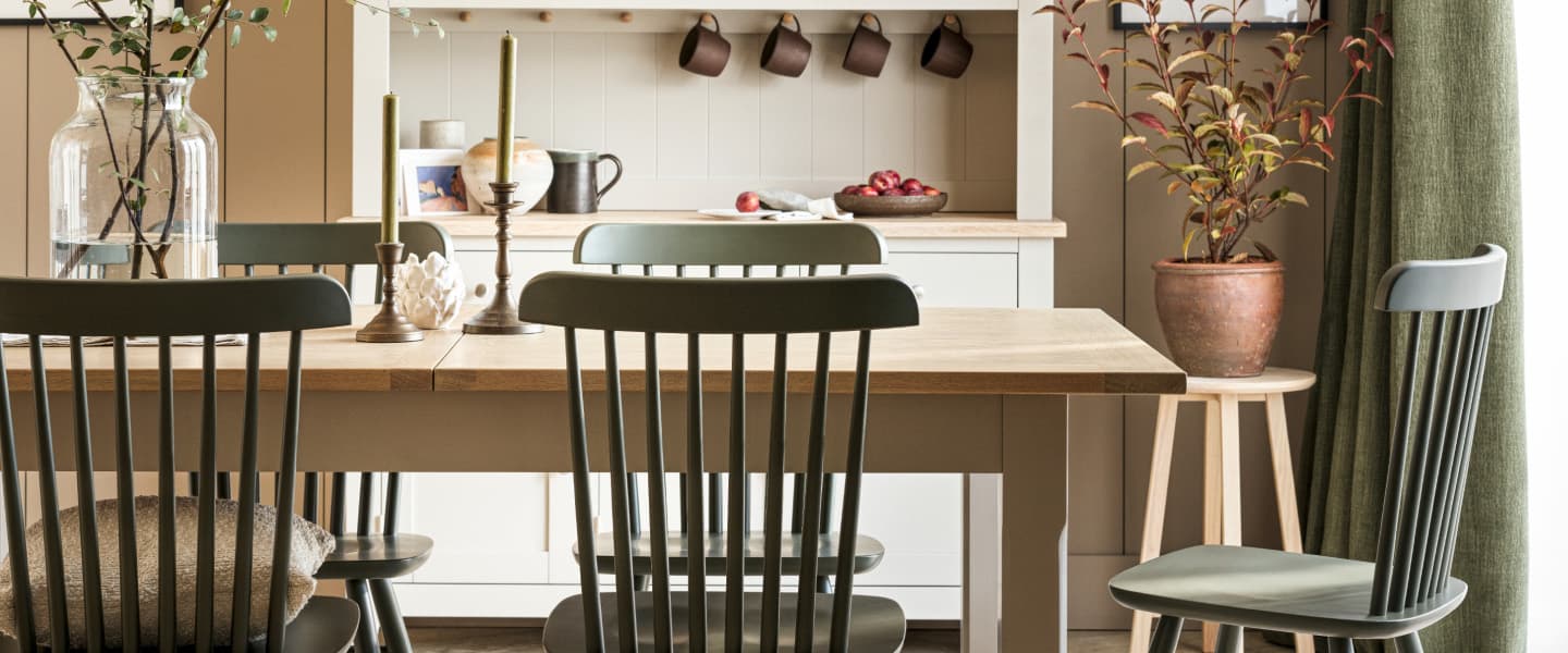 A dining room with a wooden table, green chairs, candles, a potted plant, and shelves holding cups and a bowl of apples.