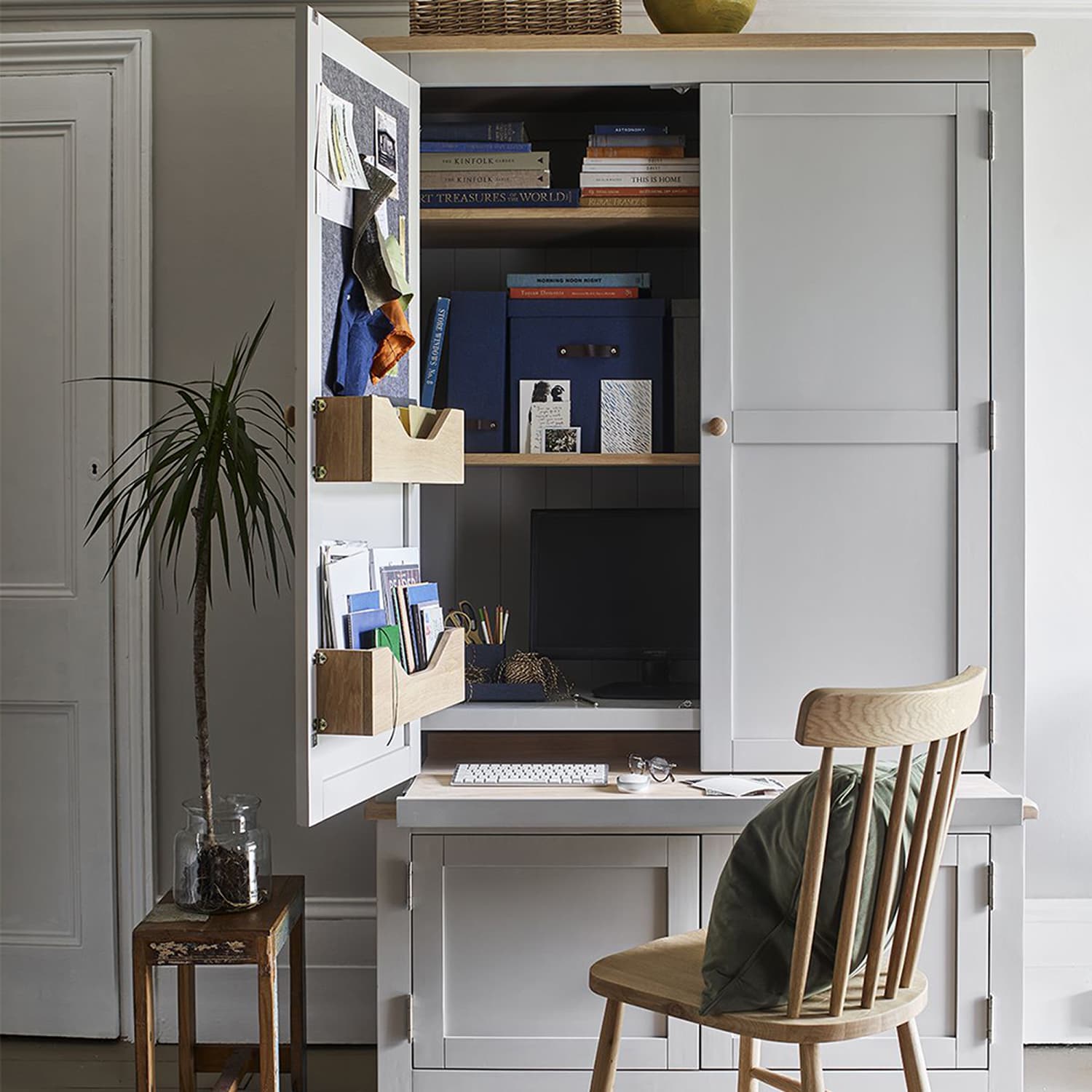 A hideaway home office unit with a wooden chair, plants, and organized shelves within an open cabinet.