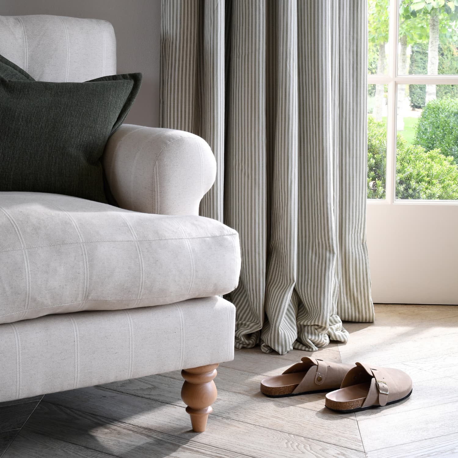Cozy room with a white armchair, dark pillow, striped curtains, and brown sandals on wooden floor near a glass door overlooking greenery.