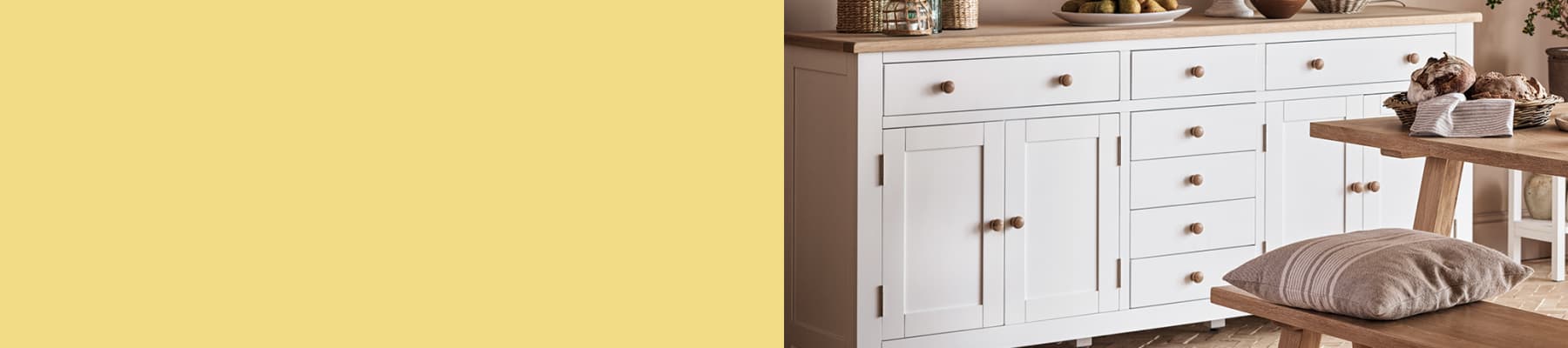White sideboard with wooden knobs, topped with decor items and fruit. Wooden table and bench in foreground.