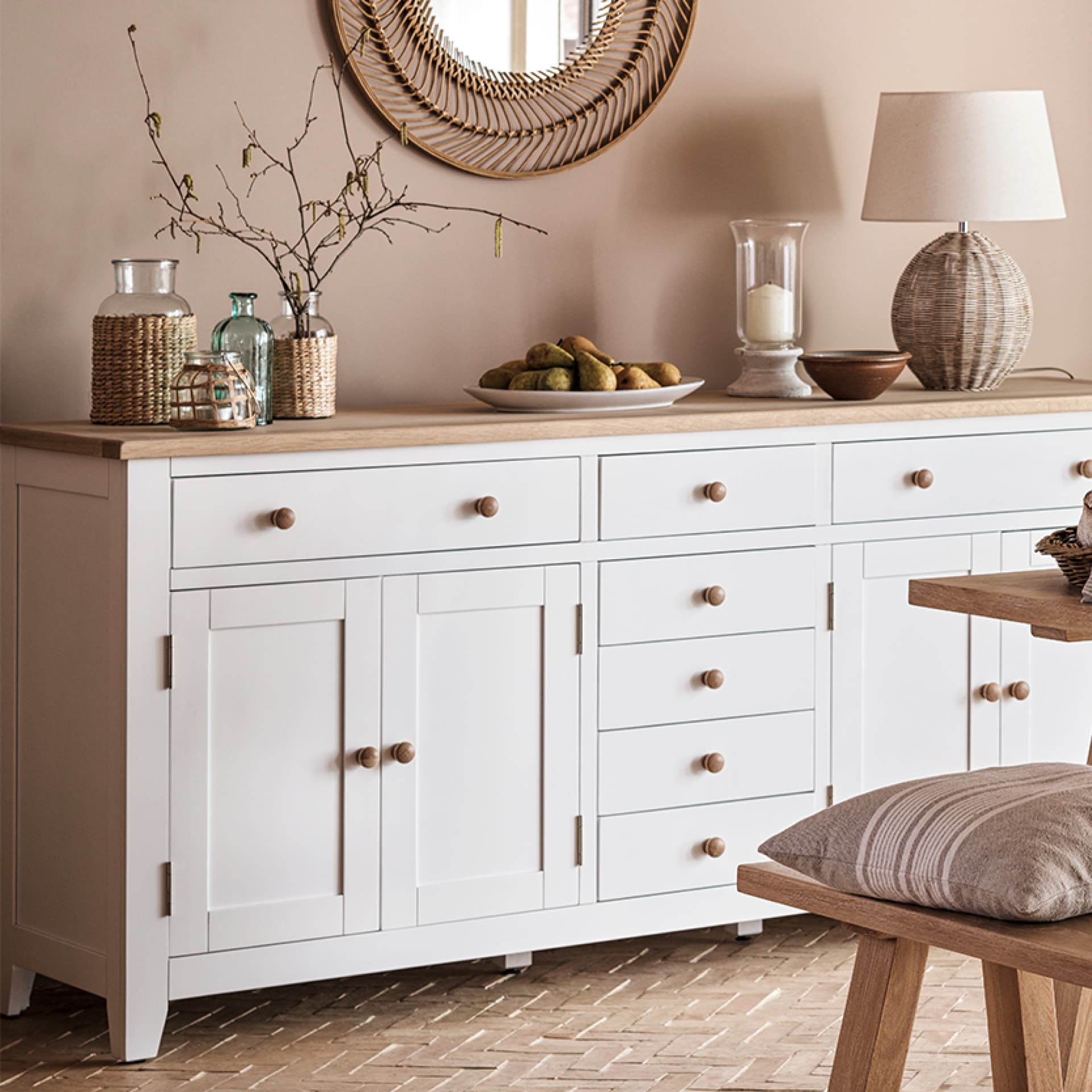 White sideboard with wooden knobs, decorated with vases, a basket, a lamp, and a round mirror above, in a warmly lit room.