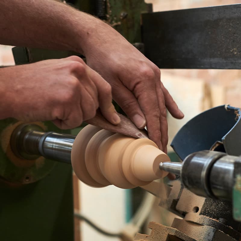 Hands shaping a spinning wooden piece on a lathe, with wood shavings visible, in a workshop setting.