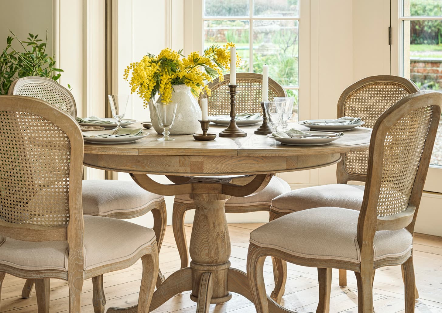 A round wooden dining table with elegant tableware, a vase of yellow flowers, and four cane-backed chairs in a bright room with large windows.