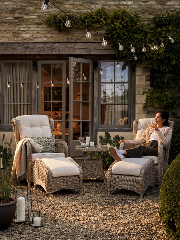 Cozy patio scene with a person relaxing on a wicker chair. Surrounding elements include cushions, fairy lights, candles, and a stone building backdrop.