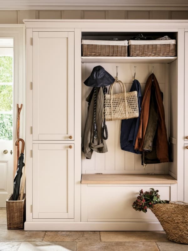 Cream-colored mudroom unit with coats, hats, and a woven bag hanging. Baskets on the upper shelf, bench below, and an umbrella stand nearby.