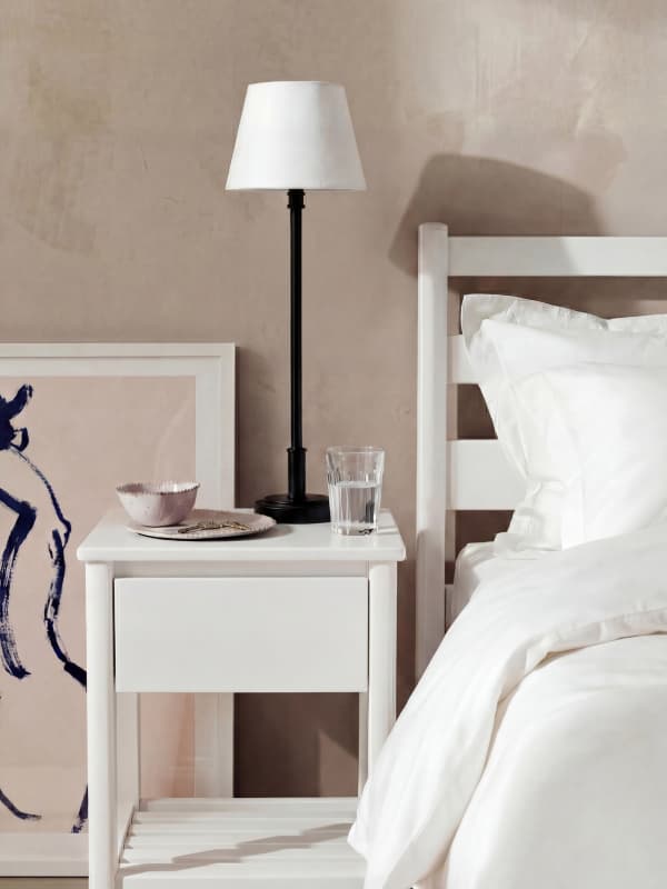 White nightstand with a lamp, glass of water, dish, and painting beside a bed with white linens against a beige wall.