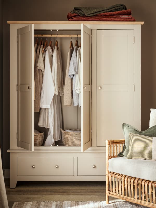 Open wardrobe with clothes, wicker baskets, and folded blankets on top. A woven chair with cushions is nearby.