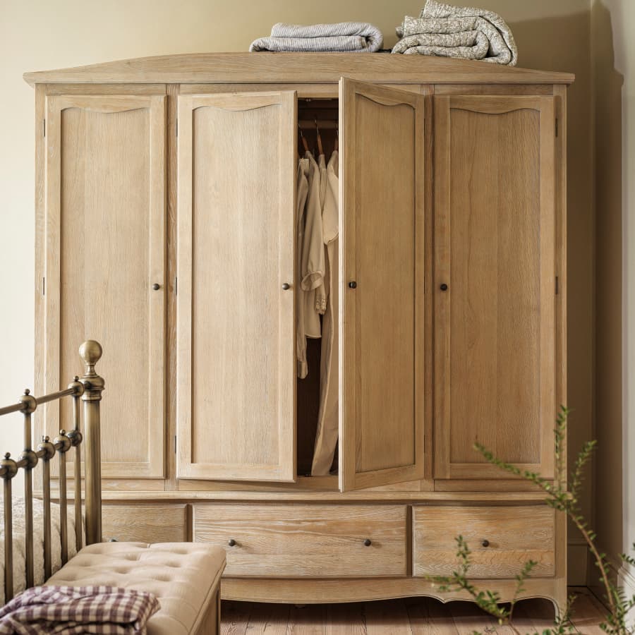 Wooden wardrobe with four doors, slightly open revealing hanging garments. Folded blankets on top, brass bed and cushioned bench nearby.